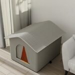 Maison isolée pour chat grise avec rabat de porte orange dans un salon lumineux à côté d’un fauteuil